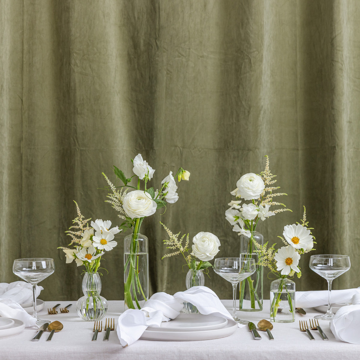 A styled dining table setting with white plates, folded white napkins, clear glass canapes and coupe glasses, and several clear glass vases holding white flowers with yellow centers.