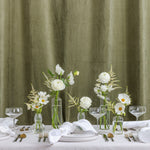A styled dining table setting with white plates, folded white napkins, clear glass canapes and coupe glasses, and several clear glass vases holding white flowers with yellow centers.