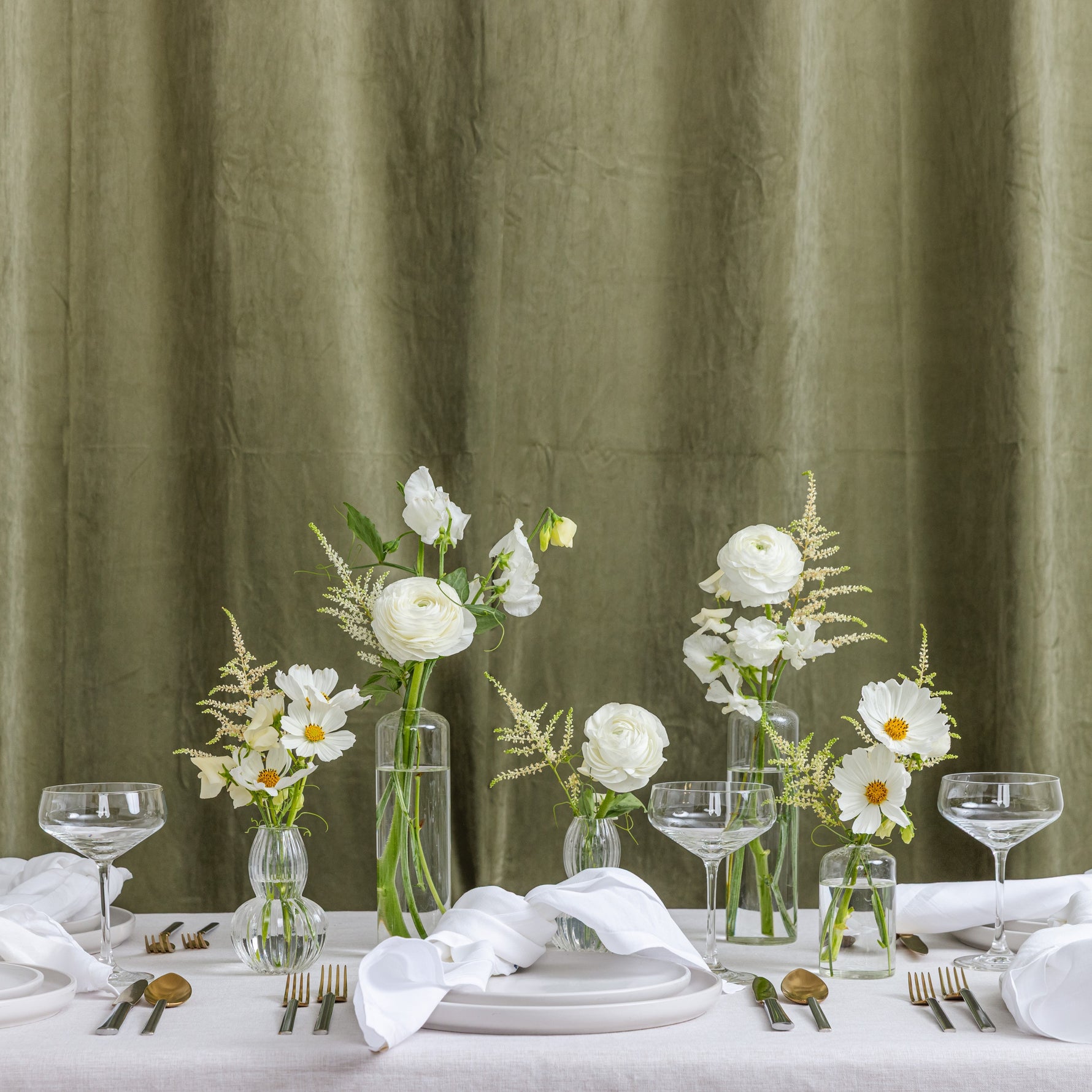 A styled dining table setting with white plates, folded white napkins, clear glass canapes and coupe glasses, and several clear glass vases holding white flowers with yellow centers.