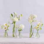 Five clear glass vases of different shapes holding white flower arrangements with green stems.