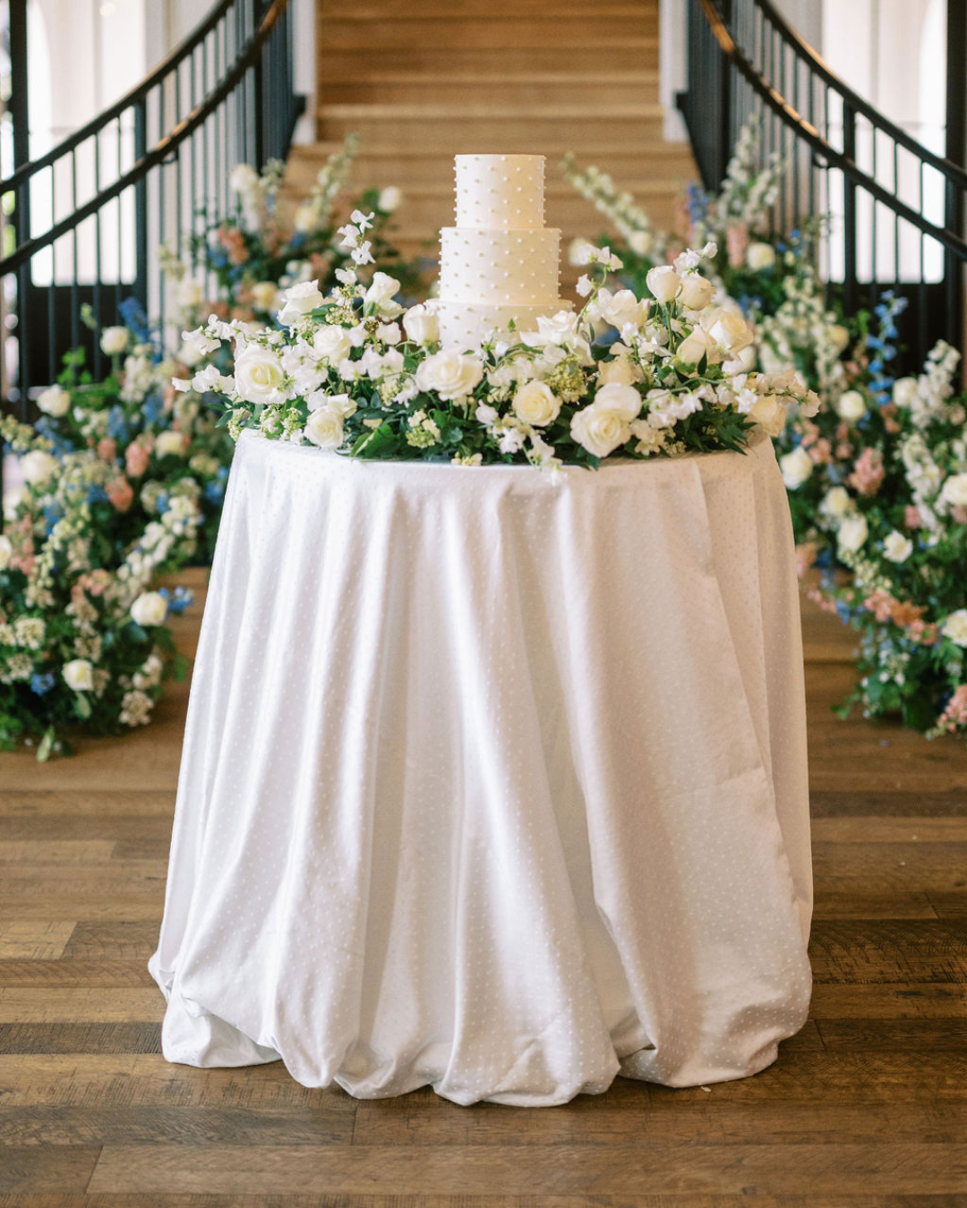 A three-tier white wedding cake sits on a round table draped in white linen, surrounded by a large floral arrangement of white and blush roses with green foliage. 