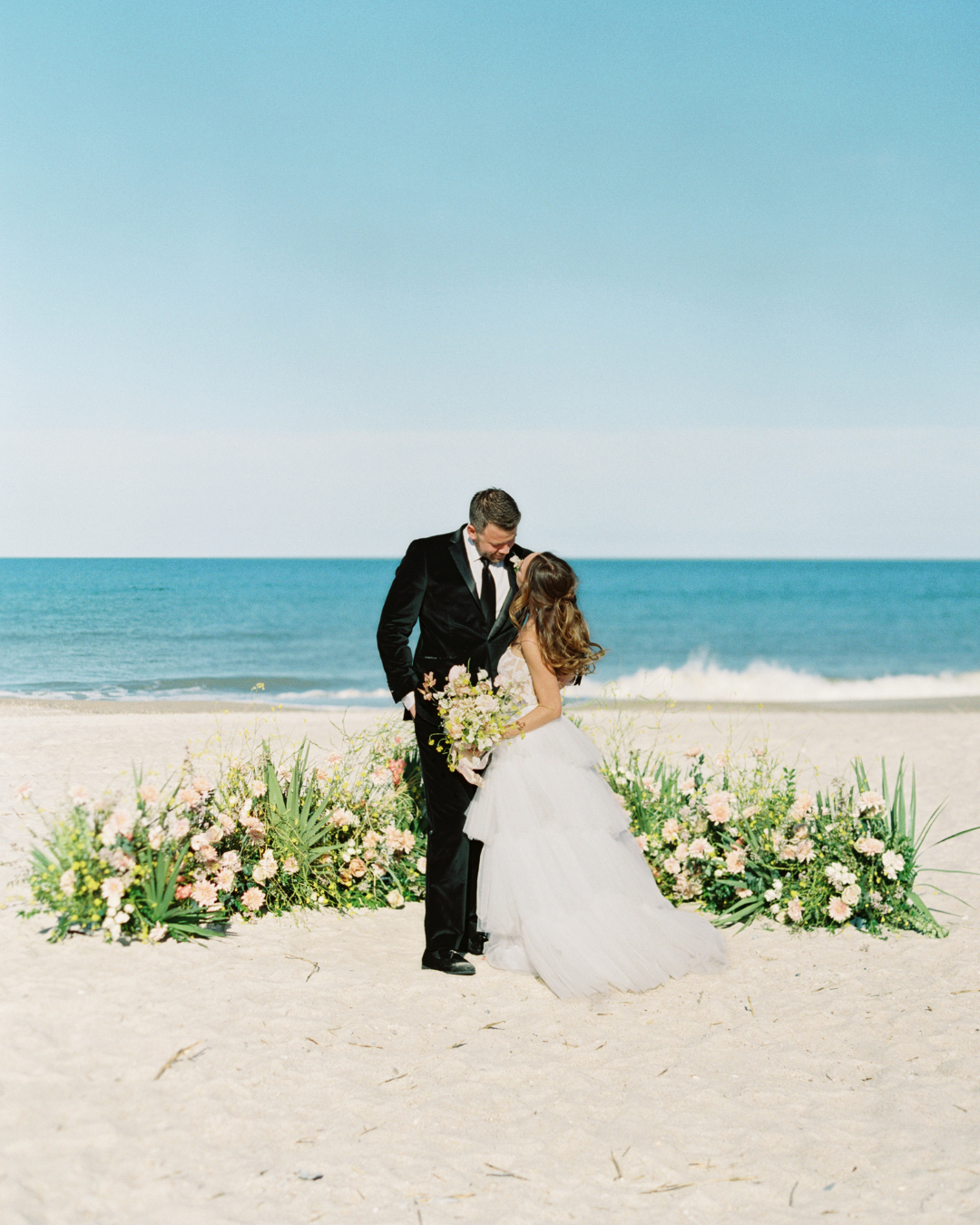 A bride in a white wedding gown and a groom in a black suit embrace on a sandy beach with a floral arch and the ocean under a clear blue sky.