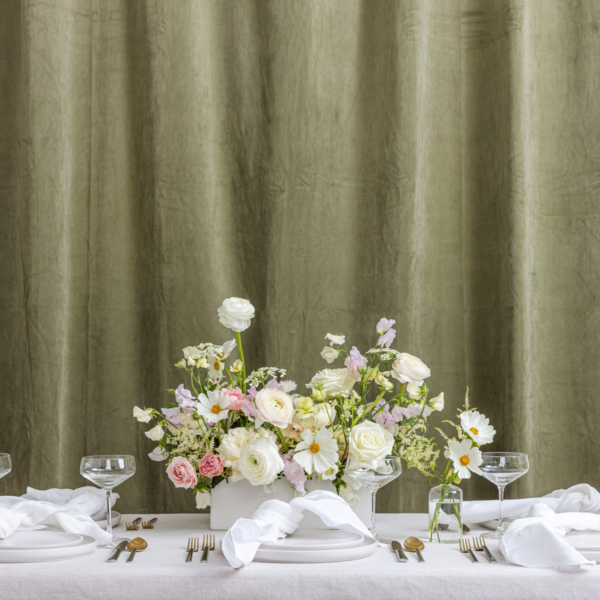 A styled wedding table set with white plates, folded napkins, silver cutlery, and wine glasses. A white vase filled with a pastel floral arrangement sits in the center.