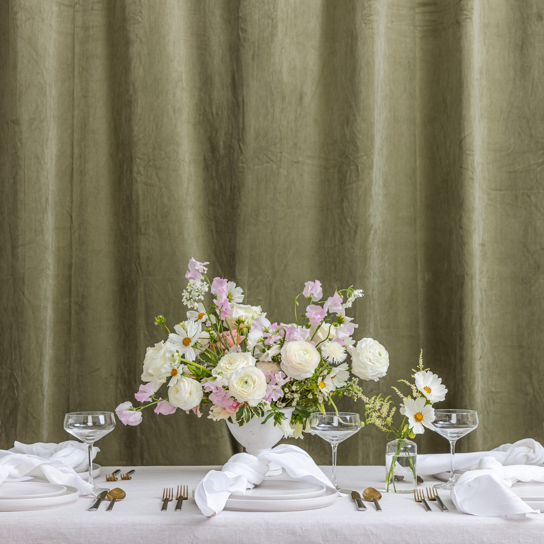 A styled wedding table setup with white tablecloths, folded napkins, silver cutlery, clear wine glasses, and a central floral arrangement in a white vase.