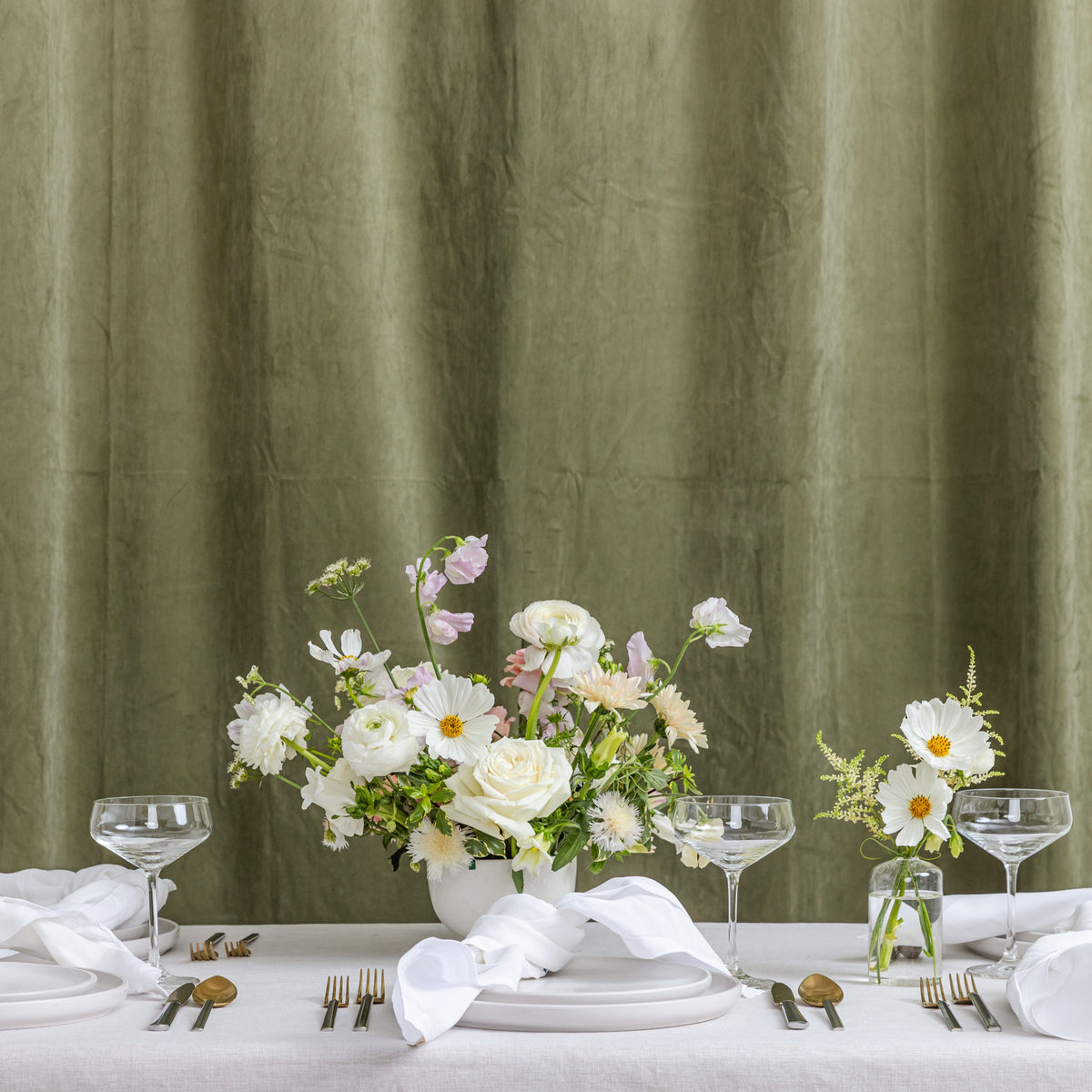 A styled wedding table setup with white tablecloths, white plates, clear wine glasses, silver cutlery, and neutral floral centerpieces.