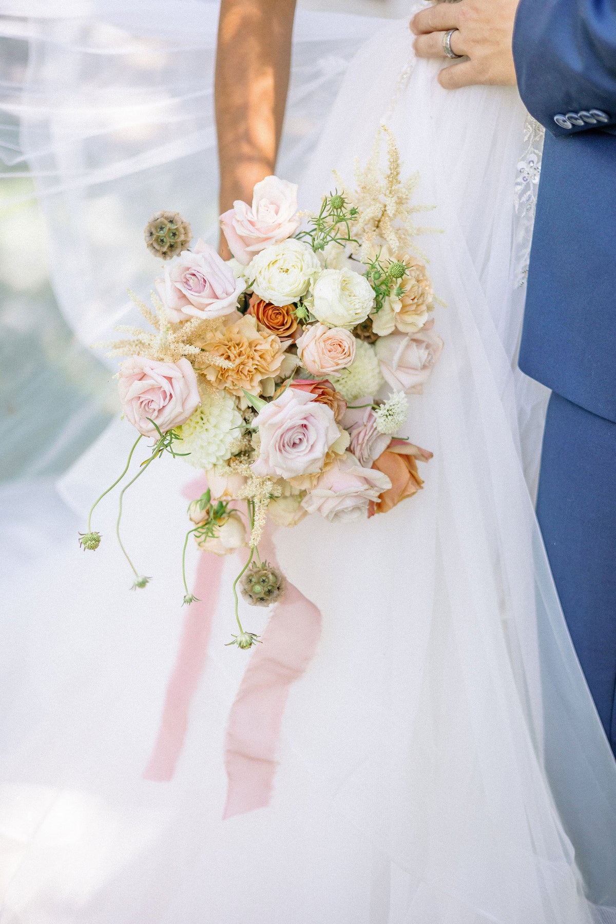 A bridal bouquet with blush pink, cream, and peach flowers, greenery, and pale pink ribbons, held against a white wedding dress.