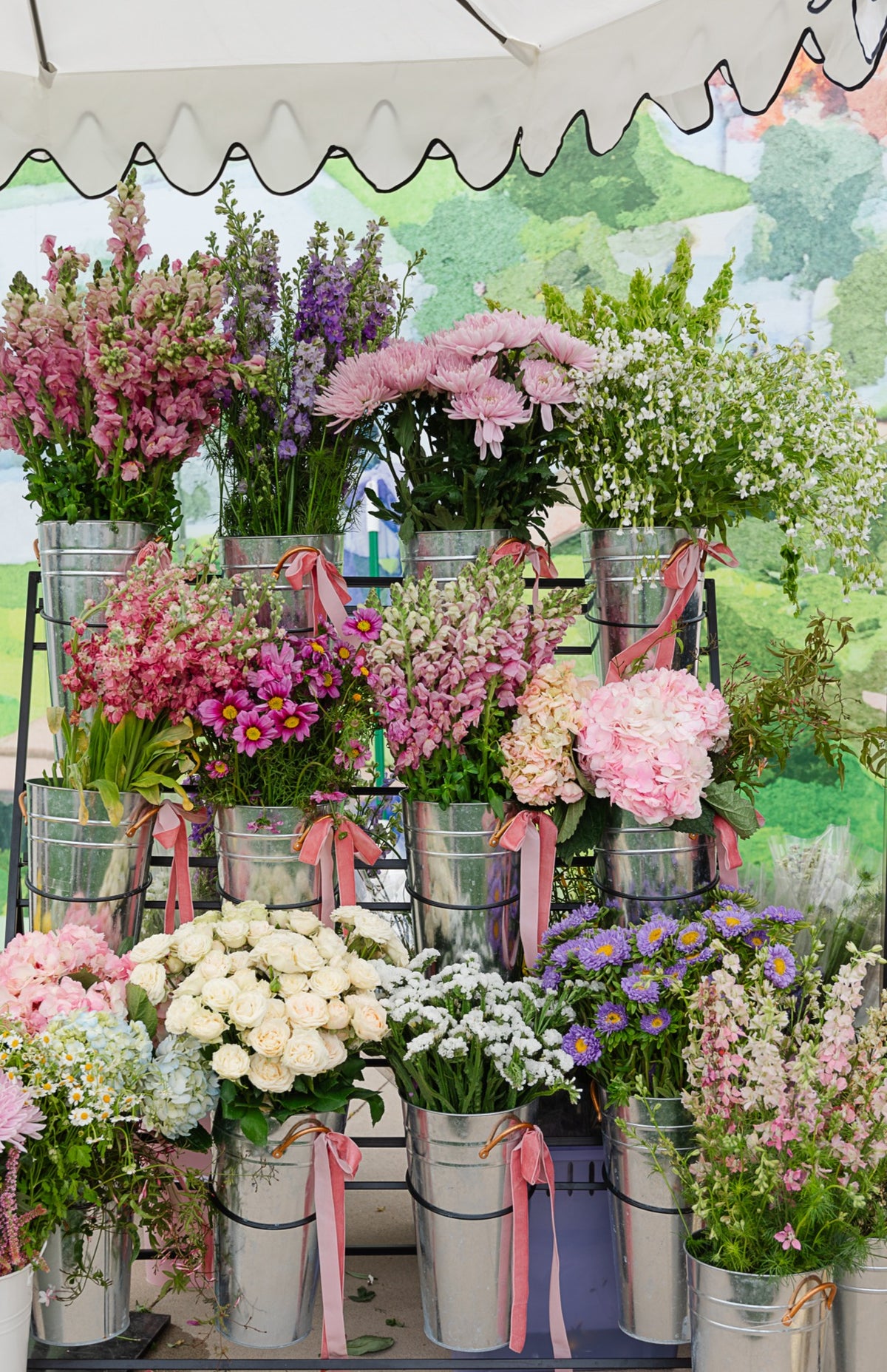 A display of assorted fresh flowers in silver metal buckets tied with pink ribbons, arranged on a stand under a white canopy. The flowers include pink, purple, white, and yellow blooms with green foliage.