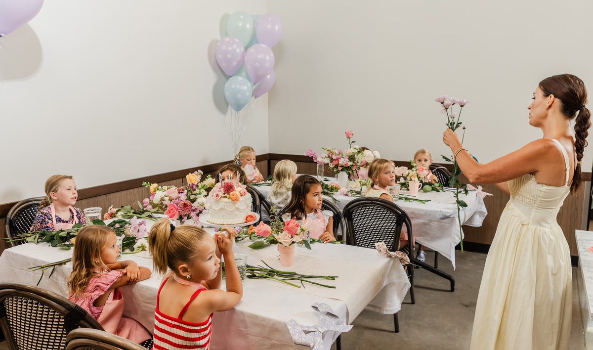 Florist Shawn Bailey stands beside a table decorated with flowers, holding a small bouquet of pink flowers while several young girls sit at the table for a flower workshop