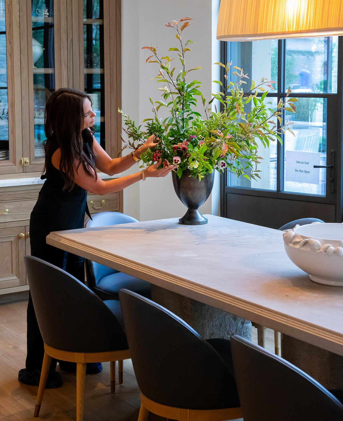 Florsit Shawn Bailey arranges a large floral arrangement in a dark metal urn on a dining table.