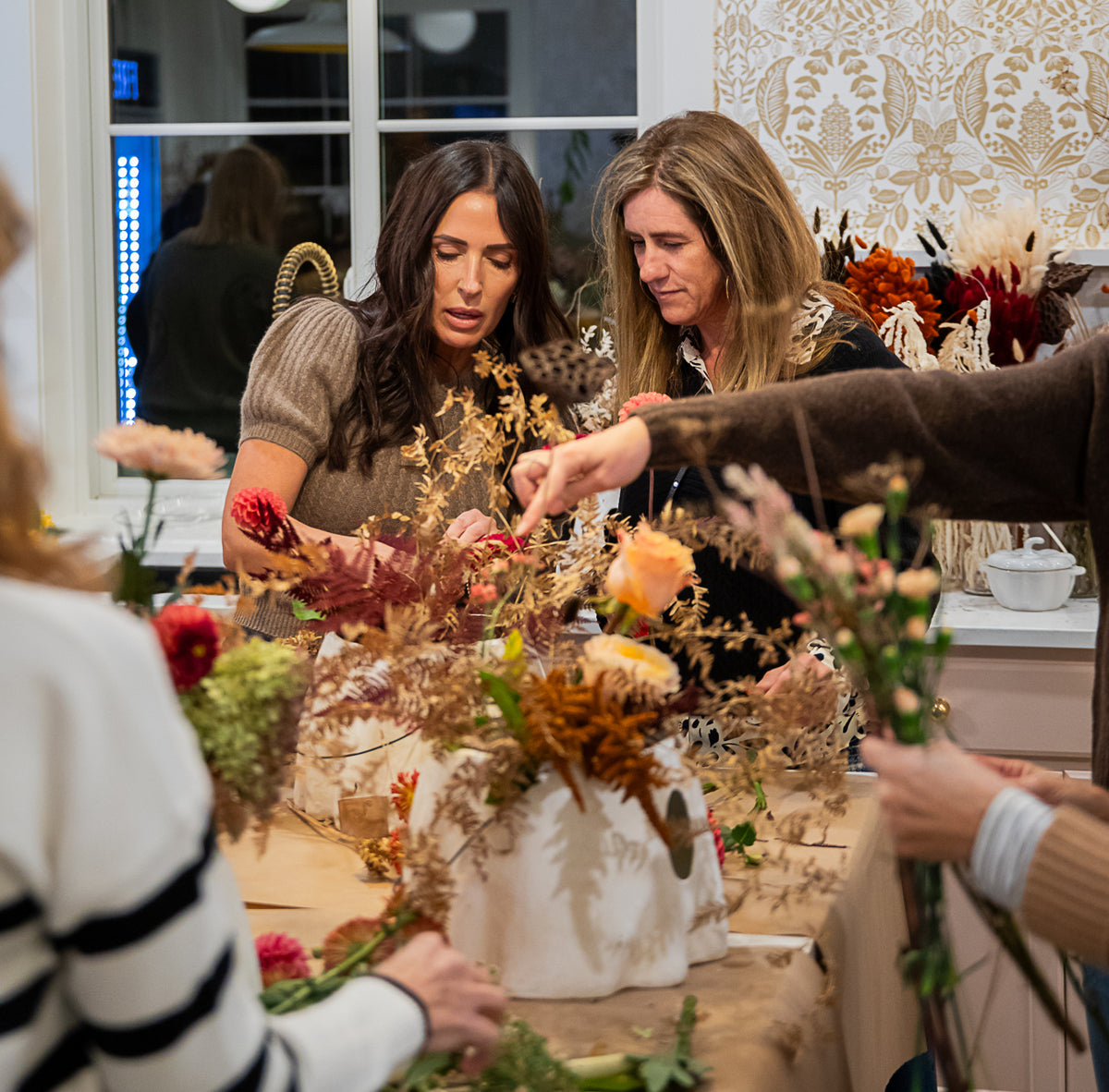 A group of women gathered around a table, arranging dried and fresh fall flowers in a ghost bucket.