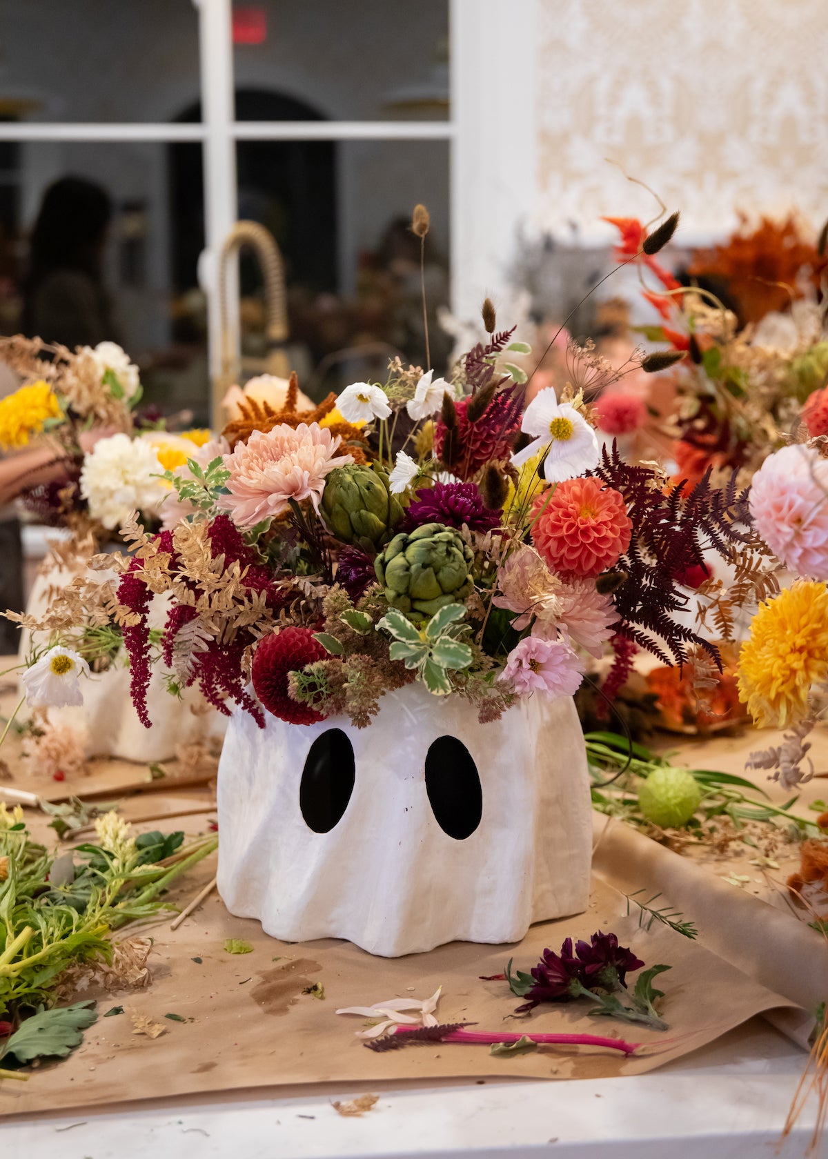 A white papier-mache ghost-shaped planter filled with a dense, colorful wildflower arrangement with artichokes and fall florals. 
