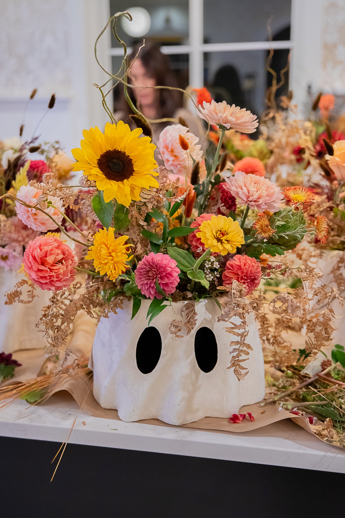 A white ghost bucket planter filled with a colorful wildflower arrangement featuring a yellow sunflower, pink and orange flowers, and green foliage.