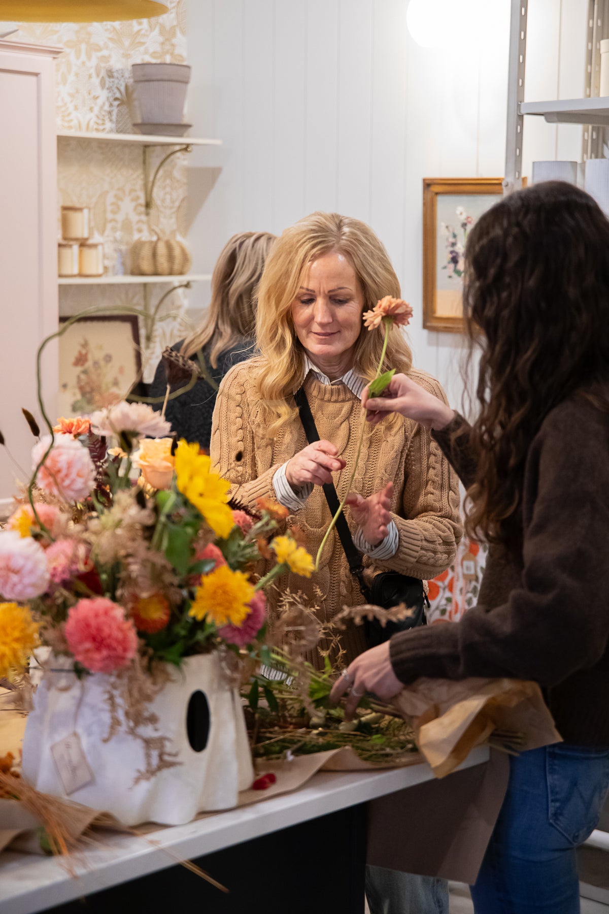 A woman stands in a flower shop holding a small orange flower during a fall floral workshop.
