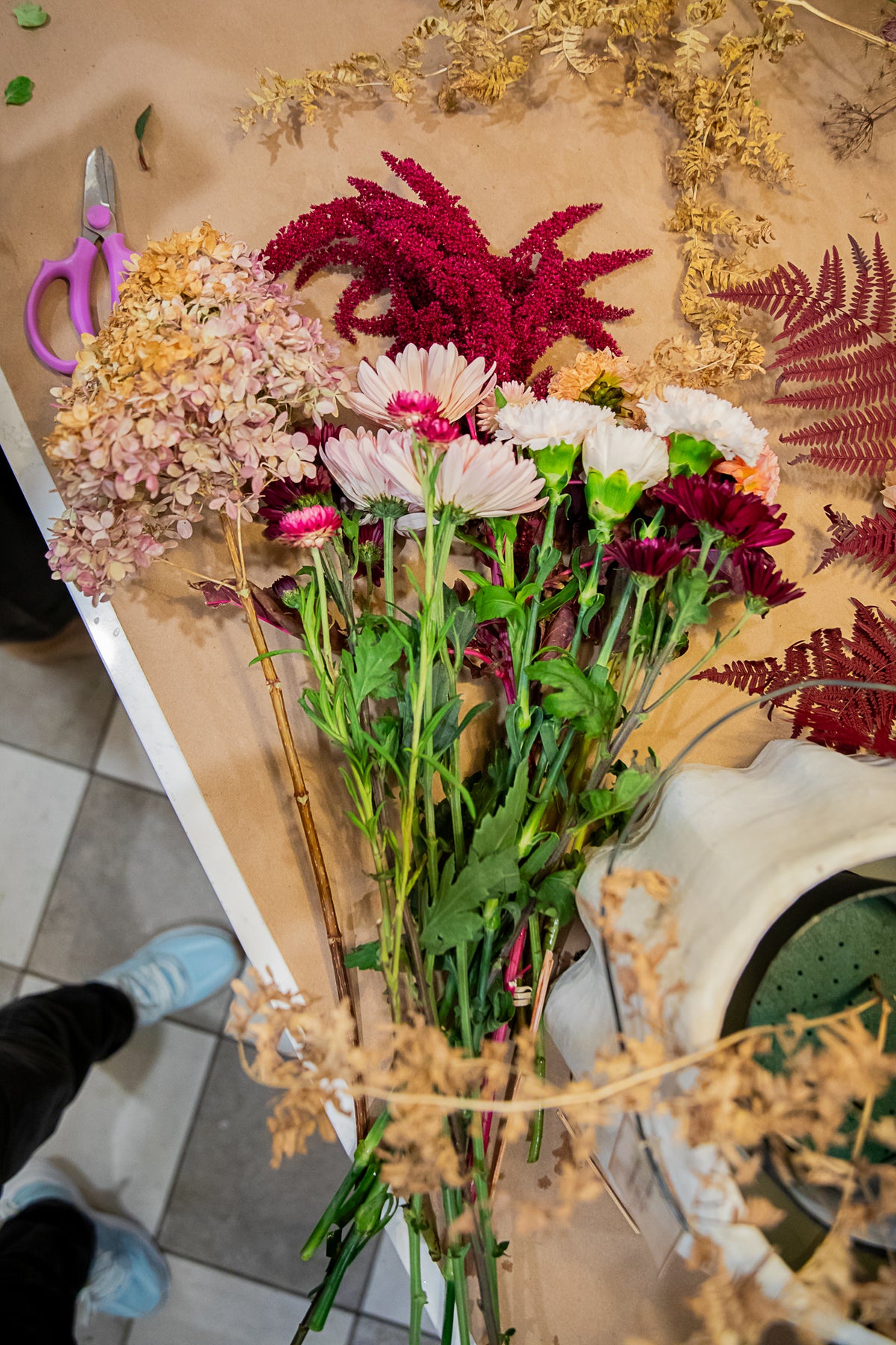 A bouquet of flowers, including pink and white daisy-like blooms, deep red feathery stems, and dried beige foliage. 