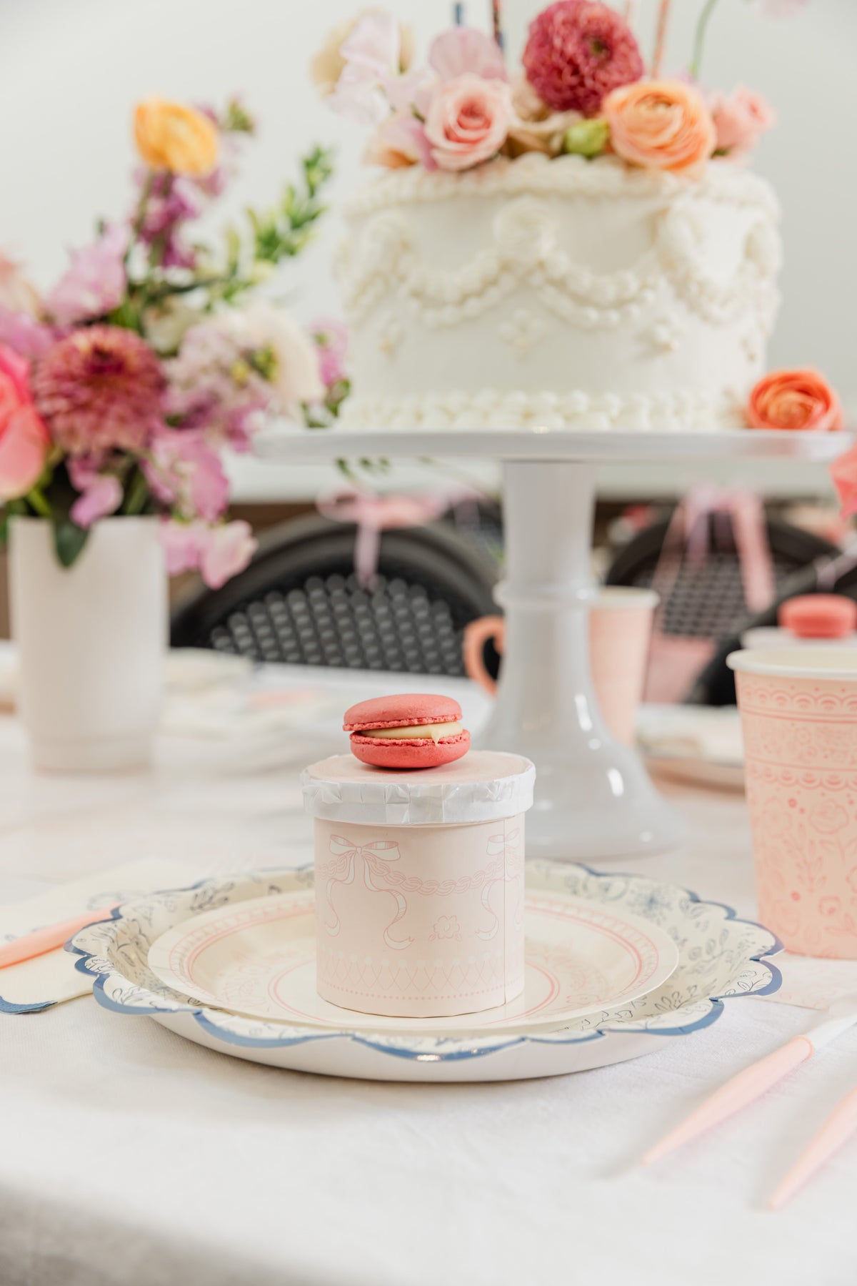 A styled tea party table scene with a white frosted cake on a pedestal stand, pink macarons, pastel cups, plates, and flowers in the background.
