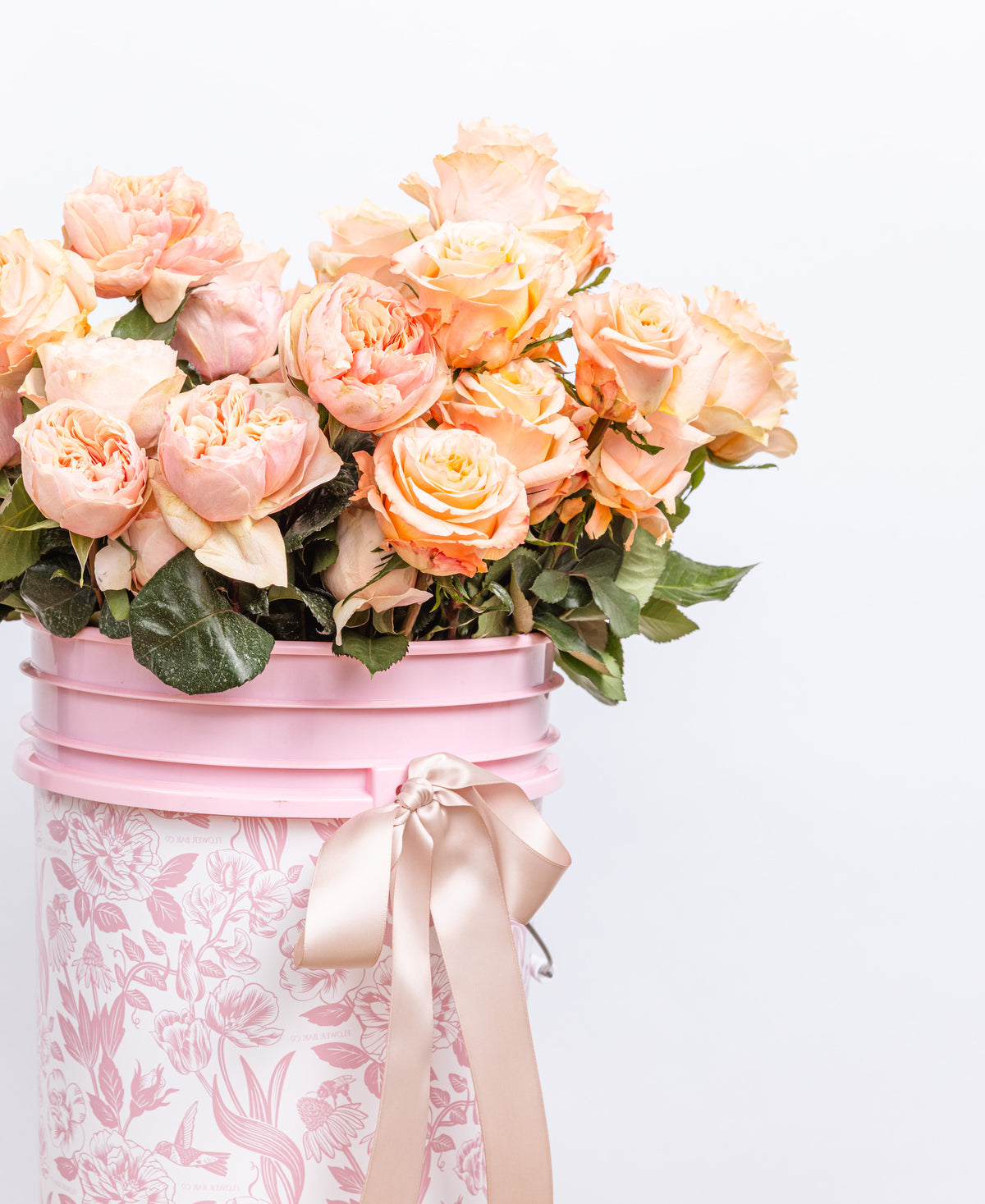 A bouquet of peach and pink roses with green leaves arranged in a pink flower bucket with a floral pattern and a satin ribbon bow.