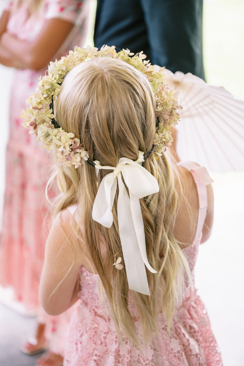 A flower girl wears a flower crown with cream and pale pink blossoms, a large white ribbon bow in her hair, and a pink dress.