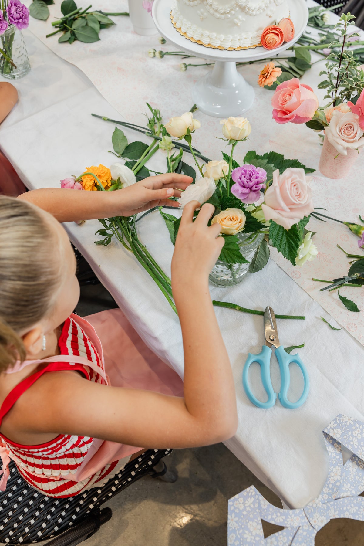 A young girl arranges a bouquet of pink, yellow, and purple roses. Blue-handled pruning shears and other flower stems are scattered on the table, with a decorated cake on a stand in the background.