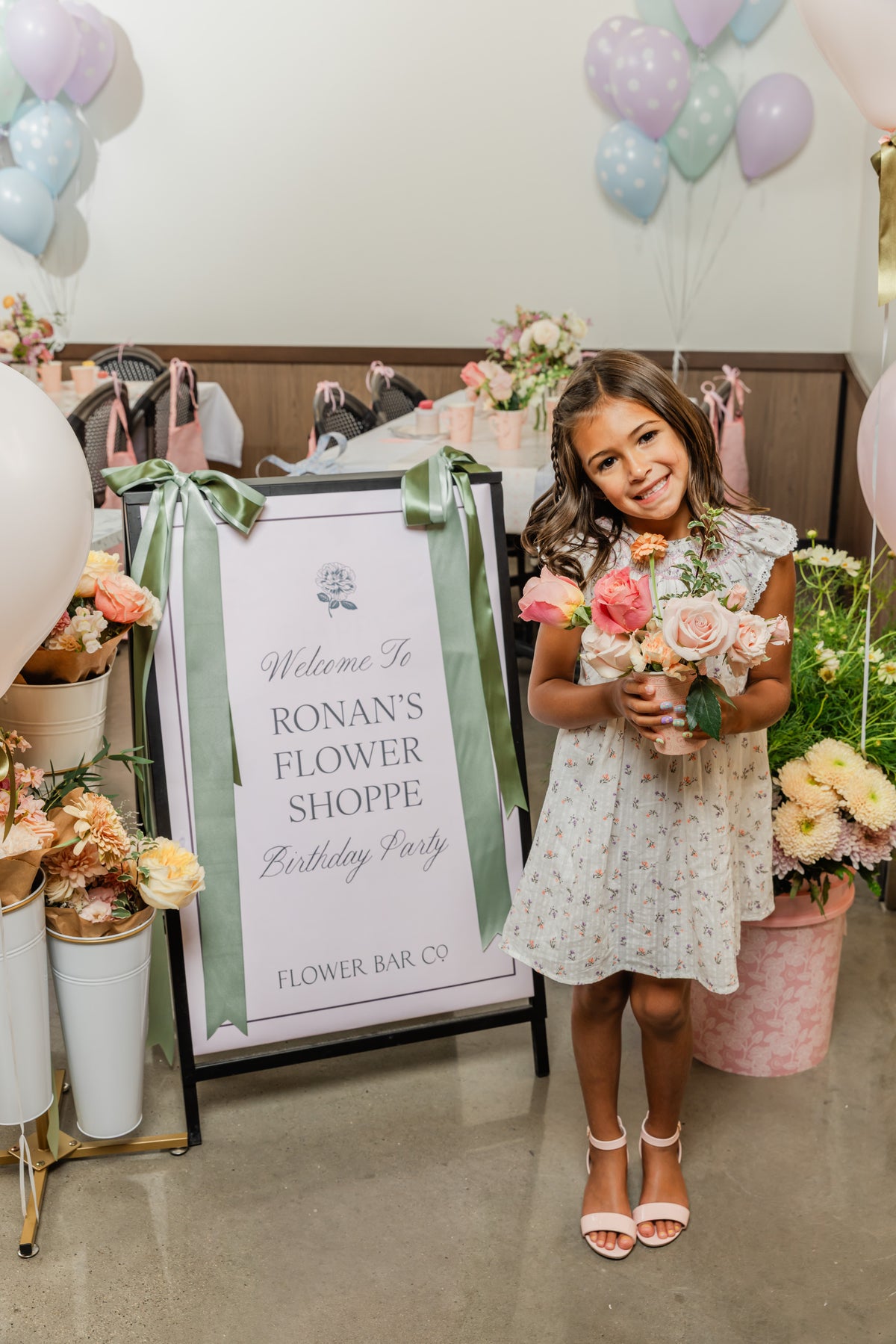 A young girl holds a bouquet of pink and peach roses next to a decorative sign that reads 'Welcome to Ronan's Flower Shoppe Birthday Party Flower Bar Co.' The birthday party is decorated with pastel balloons, flower arrangements, and party supplies.