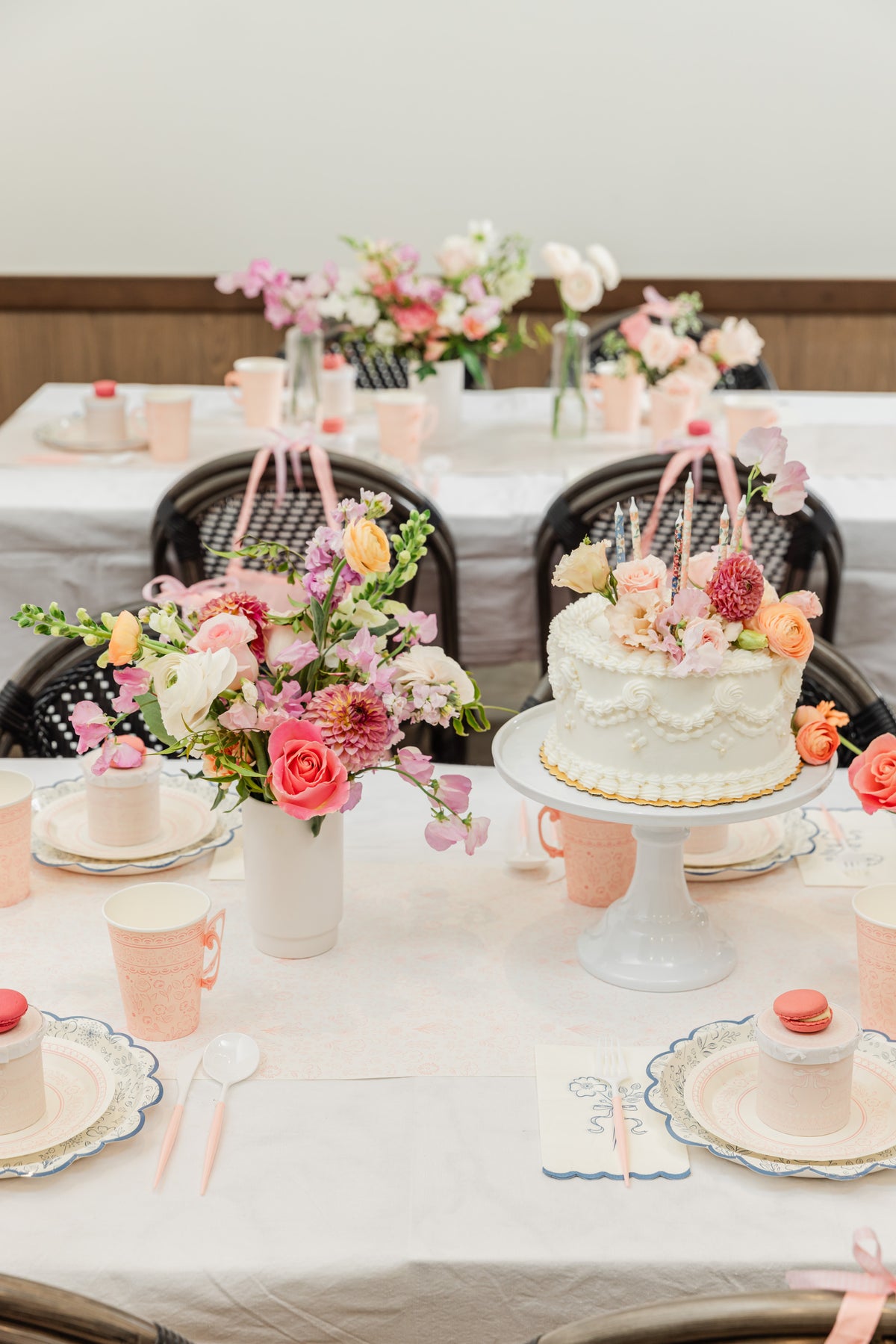 A girl's birthday party table setting with a white two-tier cake on a pedestal, surrounded by pink and peach floral arrangements, pastel pink teacups, plates, and macarons.