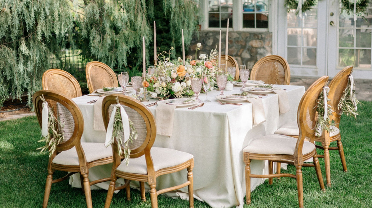An outdoor Utah wedding table setting featuring bridal floral centerpieces, tall taper candles, and wooden chairs with woven backs and cream cushions. 