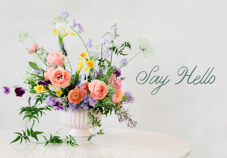 A pastel floral arrangement in a white ribbed vase with the words 'Say Hello' 