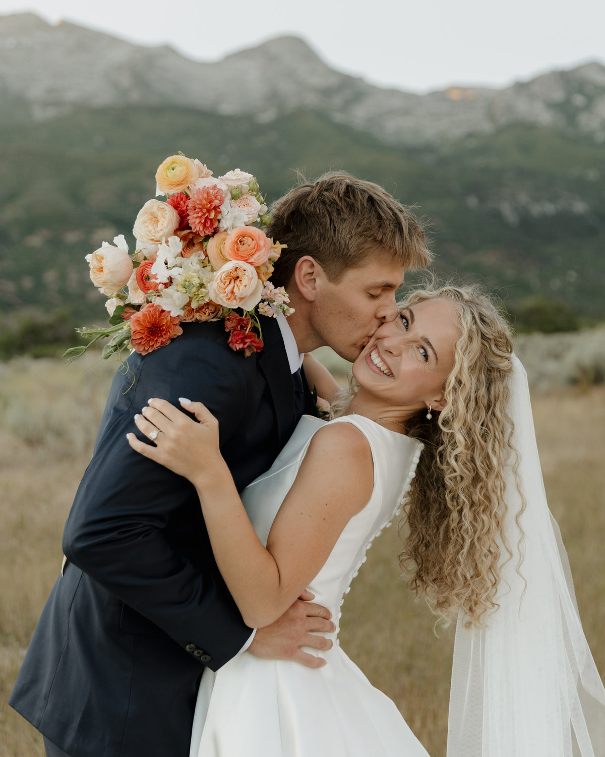 A bride kisses her groom and holds a large peach, orange, and cream floral bouquet, standing outdoors in a dry grassy field with Utah mountains in the background.